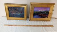 Two framed landscape paintings shown side by side on a table with a measuring stick in front, one with a forest river scene and the other with a bridge at sunset