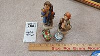 Two Hummel figurines and two small lidded boxes on carpet with ruler for size reference
