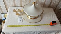 Overview of tureen with lid on plate, measuring tape for size reference, cup set shown partially