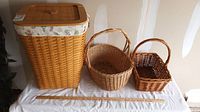Photo shows three baskets: a tall hamper with a fitted wooden lid and floral fabric lining, a large oval basket with a handle, and a smaller basket with a curved handle, all placed on a white cloth.
