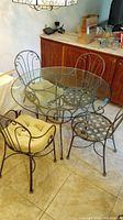 Round wrought iron dining table with glass top and four matching chairs with beige cushions arranged around it on tiled floor near wood cabinets.