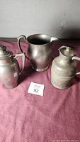Three pewter pitchers on a pink fabric background, showing general wear and patina.