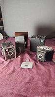 Four vintage cameras displayed on a surface; includes Ansco Shur Shot, Bell & Howell movie camera, Ansco Flash Clipper camera, and one other vintage camera with leather case.