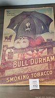 Photo of a vintage Bull Durham cardboard smoking tobacco advertising sign featuring a child under an umbrella, a small dog, and bold text branding.