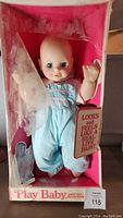 Front view of 22-inch Play Baby doll in original pink box, doll wearing blue romper, showing face and dress details.
