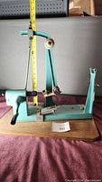 Full side view of the vintage bow maker showing the turquoise metal structure fixed on a wooden base with mechanical parts and measuring approximately 15 inches in height.