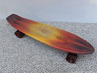 Photo of vintage 1970s skateboard deck with yellow, red, and black sunburst design and dark wheels on carpeted floor.