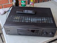 Front and top view of the Kenwood AM/FM stereo receiver 103AR showing the device with remote on top and visible ventilation slots on top surface. The unit has minor scratches and dents visible on the top.