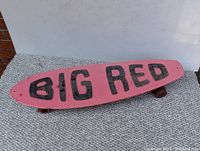 Top side view of the vintage Big Red skateboard showing pink deck and black BIG RED lettering in grip tape.