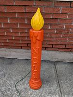 Full view of the vintage blow mold Christmas candle light standing upright outdoors against a brick wall, showing the entire orange candle base with 'NOEL' embossed and yellow flame top.