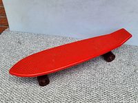 Side and angled views of vintage red plastic skateboard showing textured deck, wheels, and base with name.