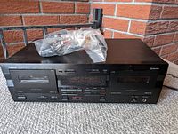Front view of Yamaha KX-W321 dual cassette deck placed on carpet with plastic bag containing remote control on top. Unit shows buttons and cassette compartments with clear Yamaha branding.