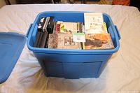 Blue plastic tote filled with various DVDs, DVD cases, and videotape cartridges on a white surface with the lid resting beside it.