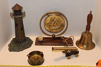 Photo showing all six brass items in the collection, arranged on white background including lighthouse statue, brass gong with ship, bell with wooden handle, ashtray, small cannon, and wooden board with mounted cannon and ball.