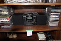 Photo showing small black CD stereo system with two black speakers surrounded by stacks of CDs on wooden shelf