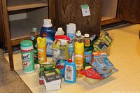 Photo showing various cleaning supplies with bottles and boxes arranged on the floor including Lysol wipes, Windex, Pledge, and bar soaps.