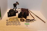 Full view of the lot including vintage coins, two paper bills, black desk clock, small sailor figurine, binoculars with leather case, belt buckles, pins, and a tube of sand.