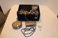 Full lot showing black container with assorted costume jewelry, blue beaded necklace, and two decorative jewelry boxes on a table.