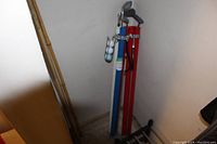 Photo showing two vintage golf caddies, one blue and one red, positioned in a corner. The blue caddy includes a metal holder for balls with multiple white golf balls inside. Several golf clubs with metal heads and black handles are seen in both caddies.