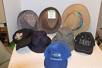 Photo showing eight hats: two patterned flat caps, a tweed flat cap, a straw sunhat with a colorful band, a purple felt fedora, a gray tweed bucket hat, and three baseball caps with logos including Seahawks, IBEW, and Vancouver Fire Dept.