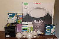 Photo showing various LED bulbs including globe bulbs, spiral CFL bulb, and standard LED bulbs in original packaging, along with Luxway Lunas LED ceiling fixture still in box.
