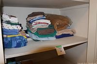 Photo showing two shelves with stacks of folded linens and towels in assorted colors. A vintage brown doormat is visible on the second shelf.