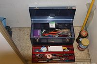 Blue metal toolbox with open lid showing assorted tools and accessories inside on a countertop with jars and broom in background