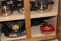 Photo of various stainless steel pots with glass lids on the top shelf and cast iron pans on the bottom shelf of a wooden cabinet. Includes a speckled Ceramic on Steel skillet with a stainless steel pot inside.