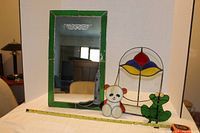 Photo of wall mirror with green stained glass frame featuring lighthouse detail, stained glass planter holder, and bear and frog animal figures in front.