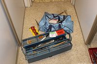 Photo showing blue metal toolbox, light blue duffel bag filled with assorted used hand tools like screwdrivers, hammer, small parts box, and a level, located on hallway floor.
