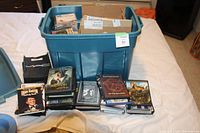 A large blue plastic tote container filled with various DVDs and CDs, some loose and some stacked in front of the container. A small MAC branded paper bag is visible.