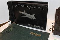 Three vintage photo albums with decorative covers: black with airplane design, green with gold text, brown with dog illustration.