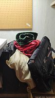 Stack of folded blankets including a green and a red one, cream-colored foam pillow, and black American Tourister bag.