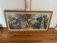 Frontal view of large horizontal framed needlepoint textile art showing three figures in 18th-century attire in a landscape with trees and a stone fountain.