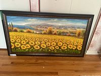 Framed acrylic painting showing a field of yellow flowers with landscape background, lying on wooden floor near wallpapered wall.