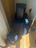 Front view of the Paradigm speaker system components including subwoofer and two satellite speakers positioned on a hardwood floor near a curtain.