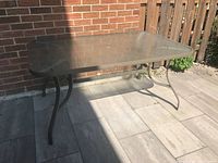 Rectangular patio table with textured glass top and curved light grey metal legs shown from side angle in bright sunlight on stone patio.
