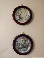 Two collector plates mounted in dark reddish wooden frames, showing traditional Asian painted scenes with figures and landscapes.
