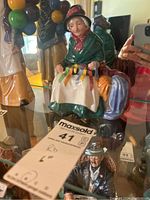 Figurine seated on a shelf with colorful ribbons visible and a MaxSold tag nearby.