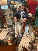 Front view of Royal Doulton figurine showing seated man holding newspaper, with clear newspaper headlines and visible wicker chair detail.