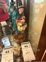 Photo showing the figurine in a glass display case with nearby items and auction labels indicating measurements and lot information.