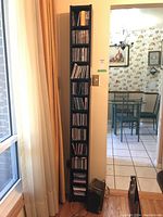 Tall slim black shelf filled with CDs positioned near a window with curtains.