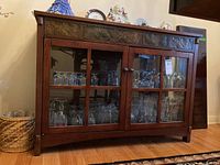 Front view of the wood sideboard showing glass doors and decorative panel above them, with various glassware inside.