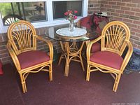 Full view of bamboo round table with glass top and two matching bamboo chairs with red cushions set on a patio.
