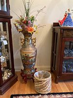 Full view of tall ceramic Asian vase on wooden stand with floral arrangement inside, adjacent woven plastic-lined basket on floor