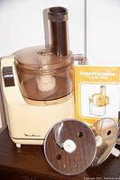 Moulinex food processor base unit with clear plastic bowl and feed chute. Stainless steel slicing disc, shredding disc, and metal blade visible. Instruction manual included in background.