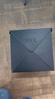 Top and front view of black metal mailbox on wooden floor with embossed 'MAIL' and some paint imperfections.