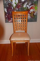 Single wooden chair with lattice back and protective cover on the padded seat, shown front-facing in a room with hardwood floor and abstract painting in the background.