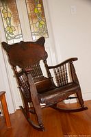 Photo of a dark wooden rocking chair with decorative carved backrest and turned spindle supports on the sides, standing on a hardwood floor.