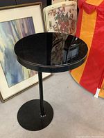 Round black glass accent table standing on an offset metal pedestal base with a circular base plate. Reflective surface and nearby framed artwork shown.